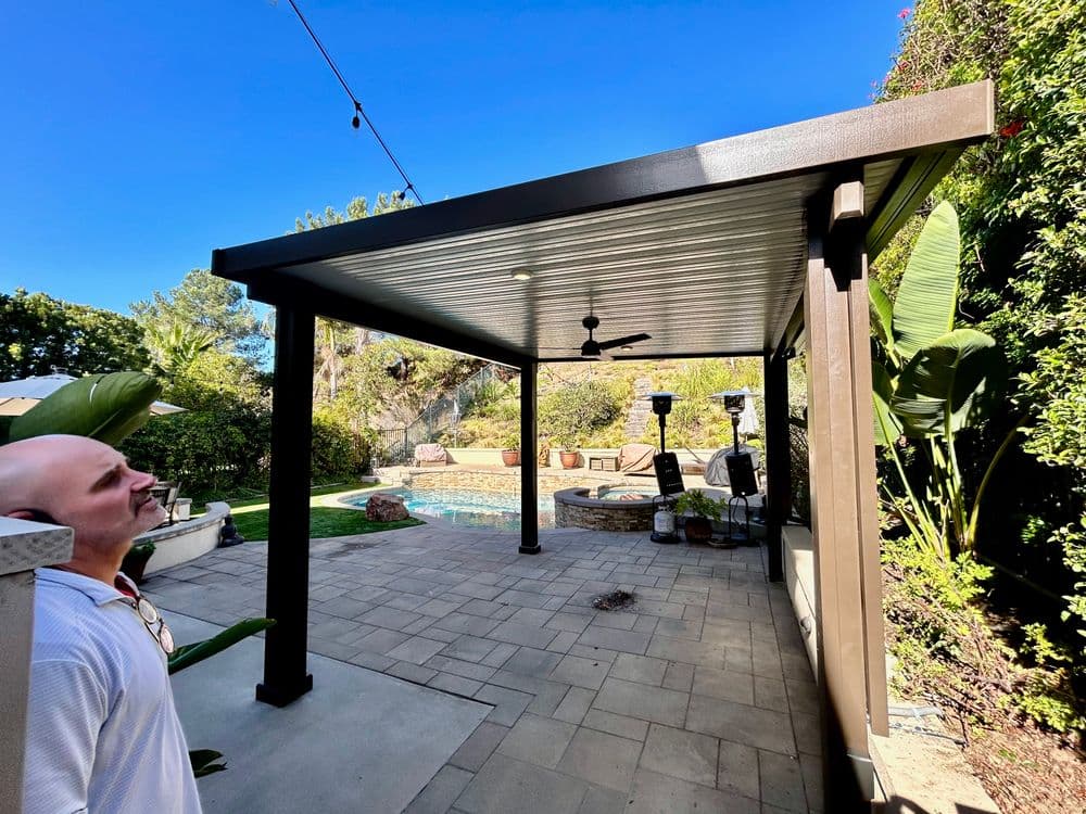 Modern backyard patio with pool, ceiling fan, and lush landscaping under a clear blue sky.
